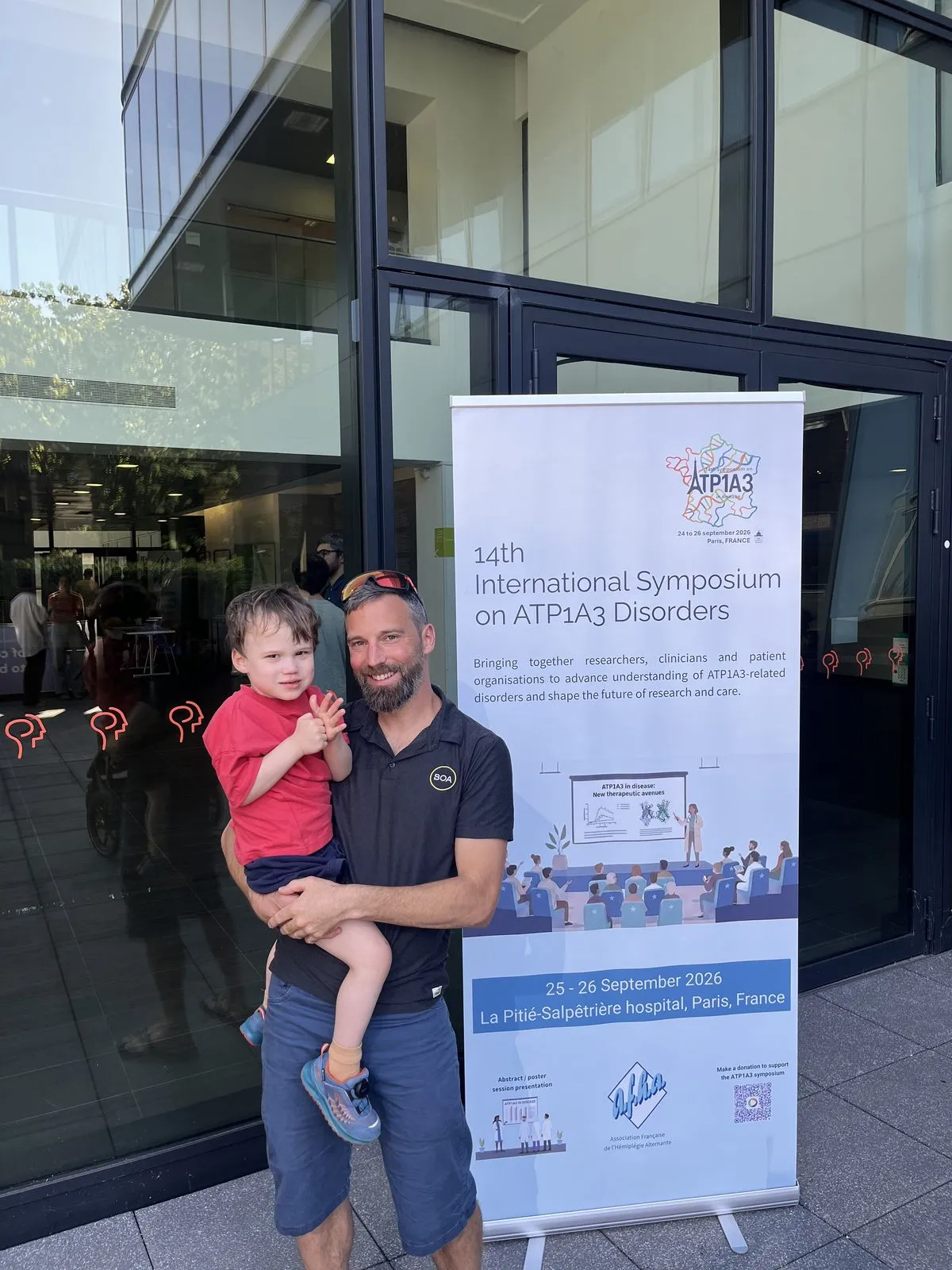 Michael with child in front of the official 14th Symposium on ATP1A3 Disorders sign in Paris